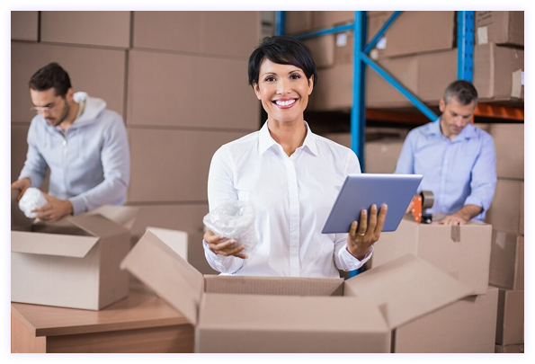 Woman in warehouse smiling with laptop and workers in background